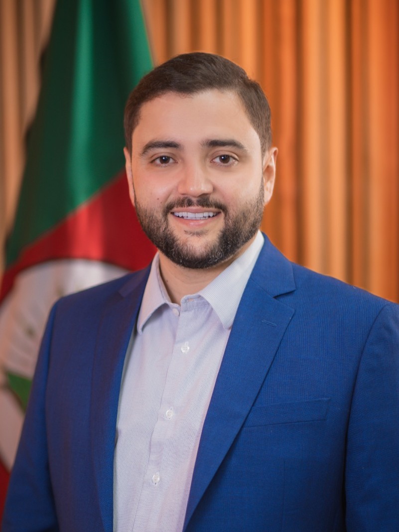 Retrato do vice-governador Gabriel Souza. Ele &eacute; um homem branco, de barba, cabelo curto. Vestindo camisa branca e blazer azul. Ao fundo, bandeira do RS.